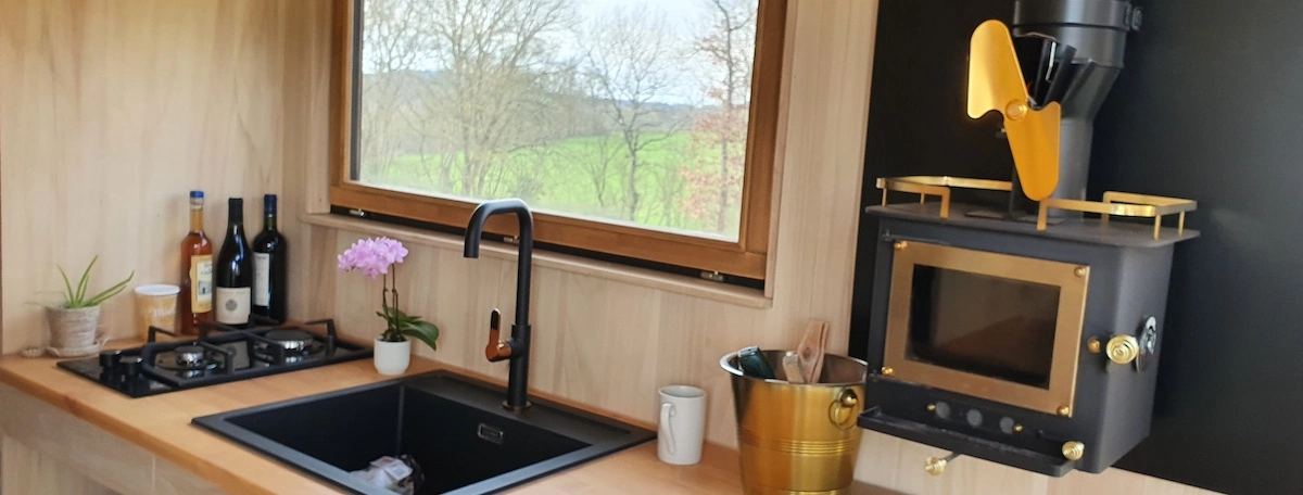Poêle à bois Cubic Grizzly Mini doré installé à mi-hauteur sur le plan de travail de la cuisine de la tiny house Youca, avec ventilateur mécanique et vue sur la campagne